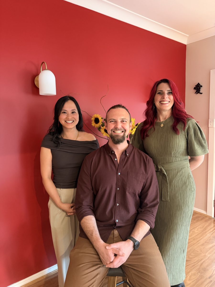 IMG_3459 Three adults smile in front of a red wall, with sunflowers in a vase behind them. One is seated and two are standing. Mornington Chinese Medicine