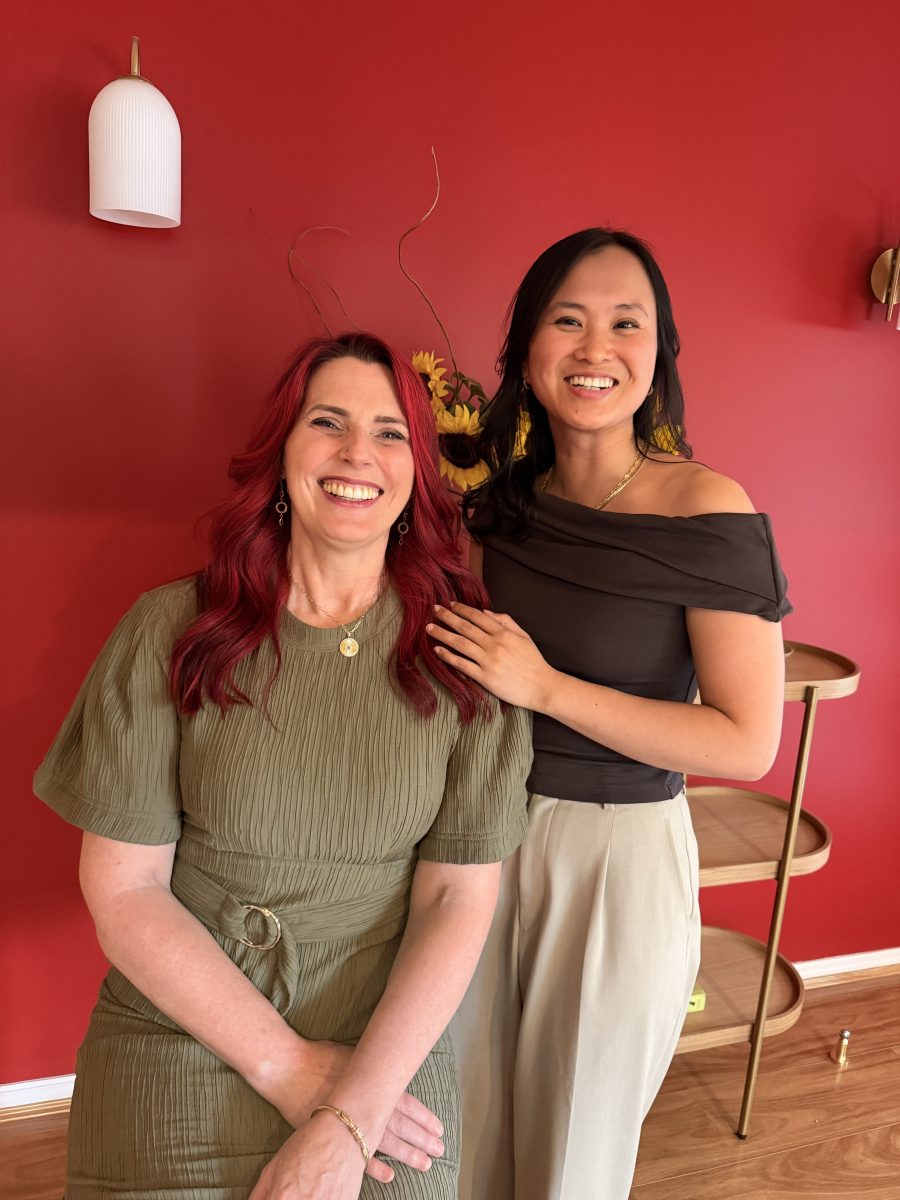 IMG_3464 Two women smiling in front of a red wall; one is seated, the other standing, both dressed in casual clothes. Mornington Chinese Medicine