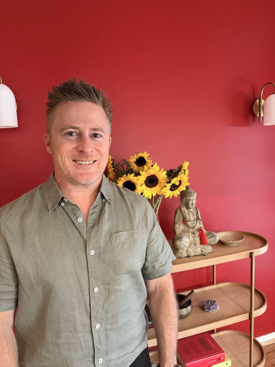 IMG_3491 Smiling man stands beside a wooden shelf with sunflowers, a Buddha statue, and books against a red wall. Mornington Chinese Medicine