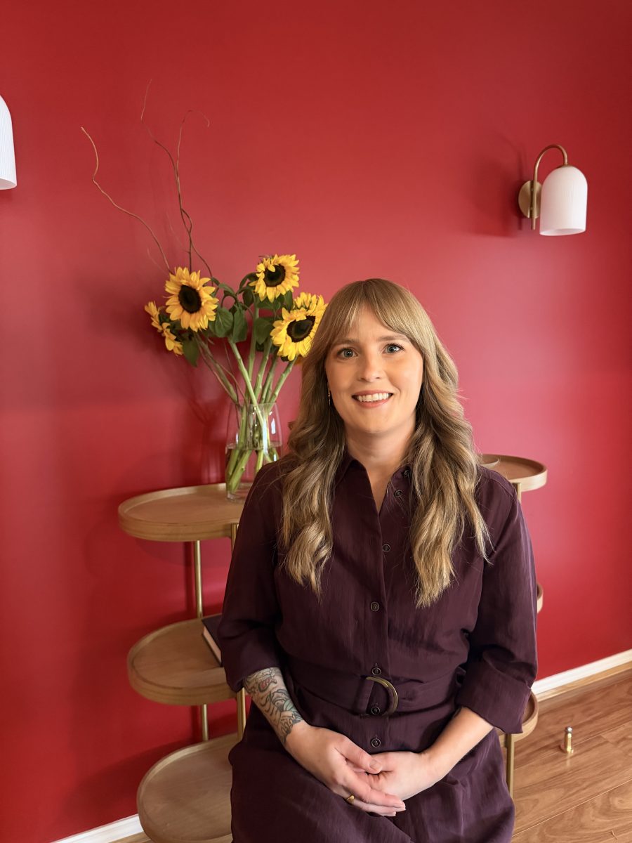 IMG_3566 A woman with long blonde hair smiles while seated before a red wall, with sunflowers in a vase on a wooden table behind her. Mornington Chinese Medicine