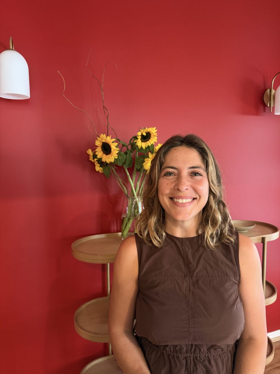IMG_3576 Smiling woman in a brown dress stands before a red wall, with sunflowers in a vase and wooden shelves visible behind her. Mornington Chinese Medicine