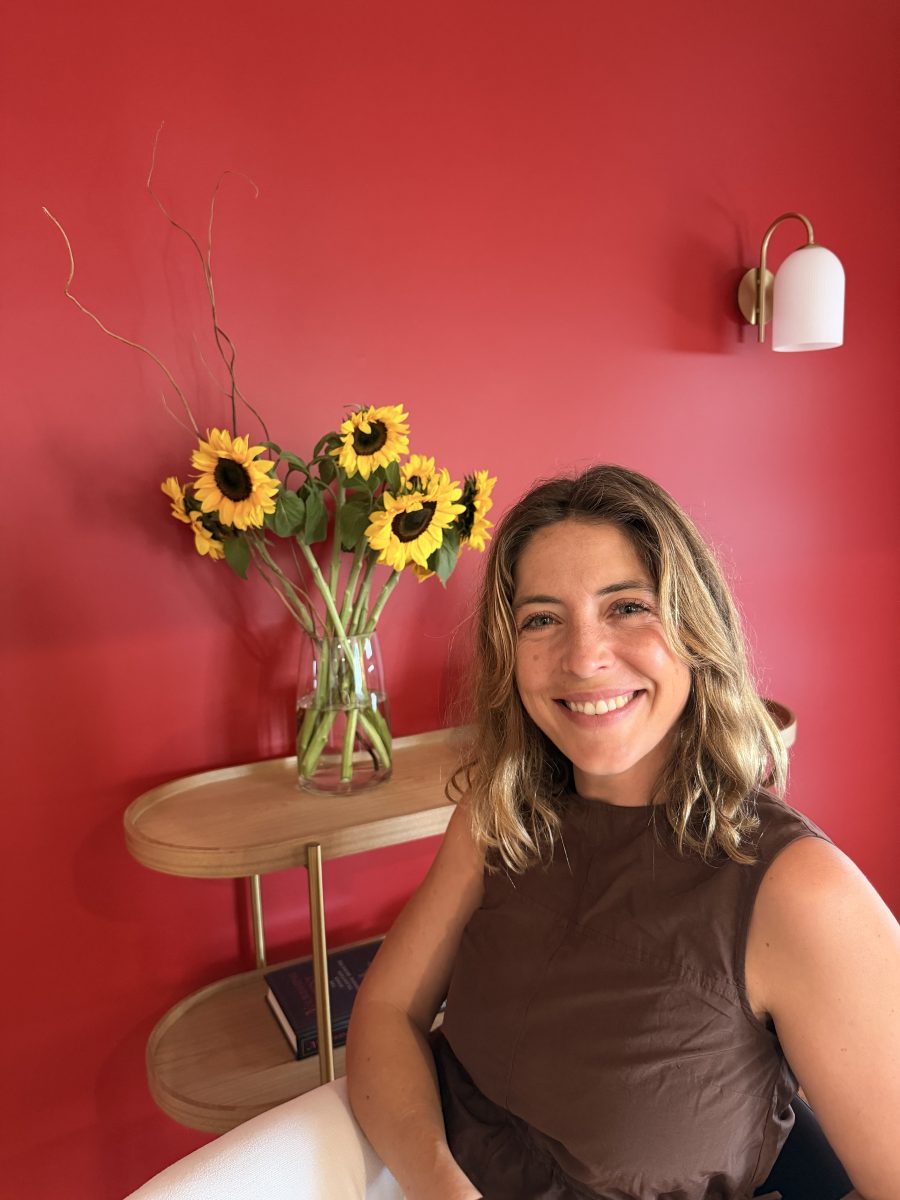 IMG_3582 Smiling woman with wavy hair sits before a vase of sunflowers, set against a red wall featuring a modern wall lamp. Mornington Chinese Medicine