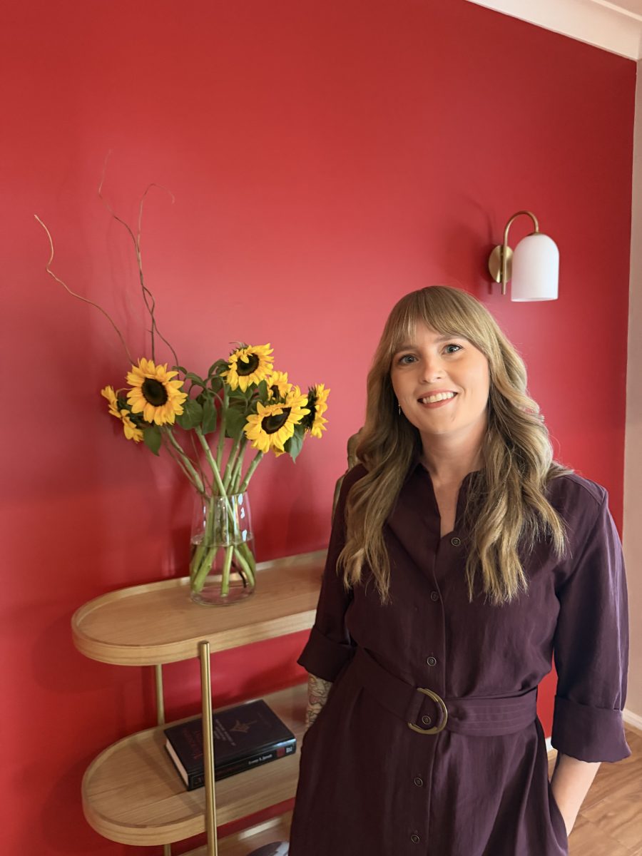 IMG_3638 Woman in a purple dress stands by a wooden table with a vase of sunflowers, set against a red wall. Mornington Chinese Medicine