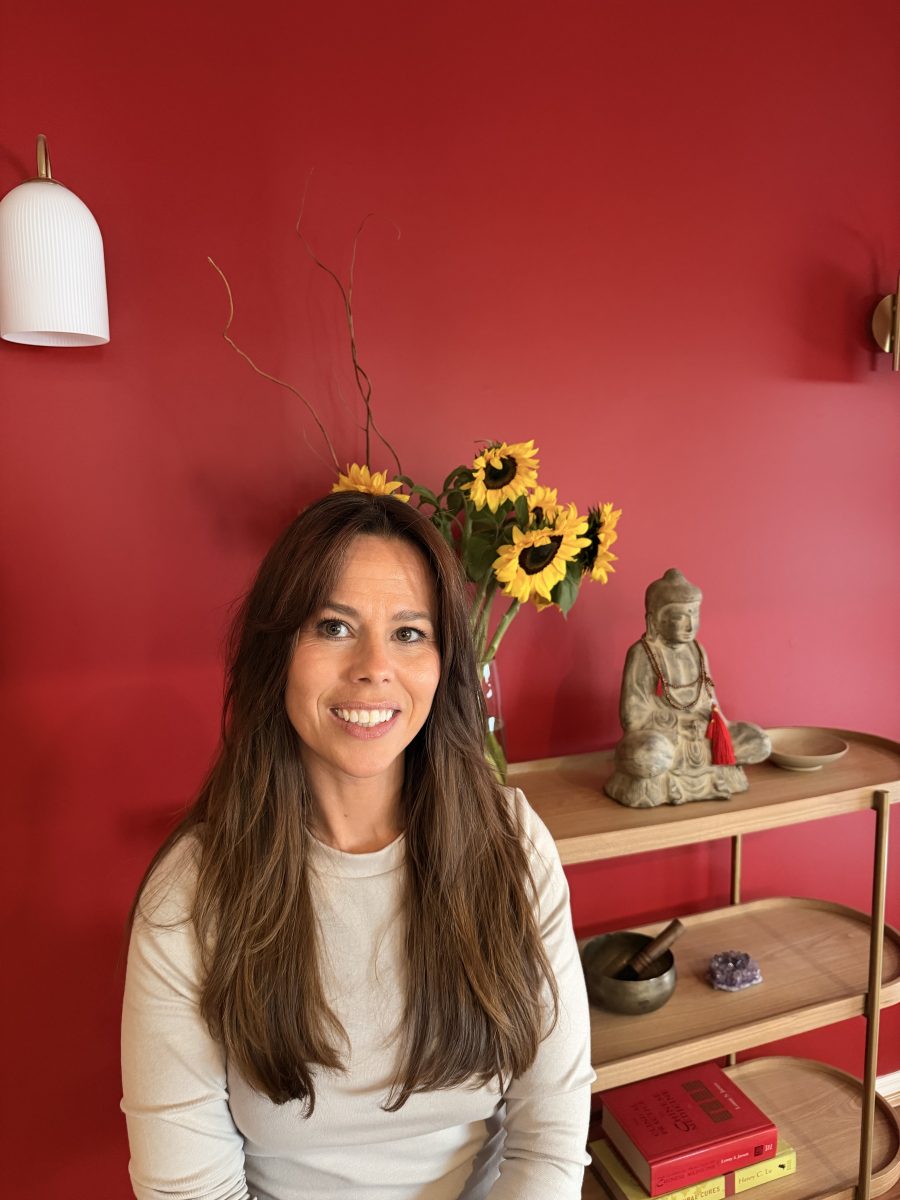 IMG_3672 Woman with long brown hair smiles before a red wall, sunflowers, and a shelf holding a Buddha statue and books. Mornington Chinese Medicine