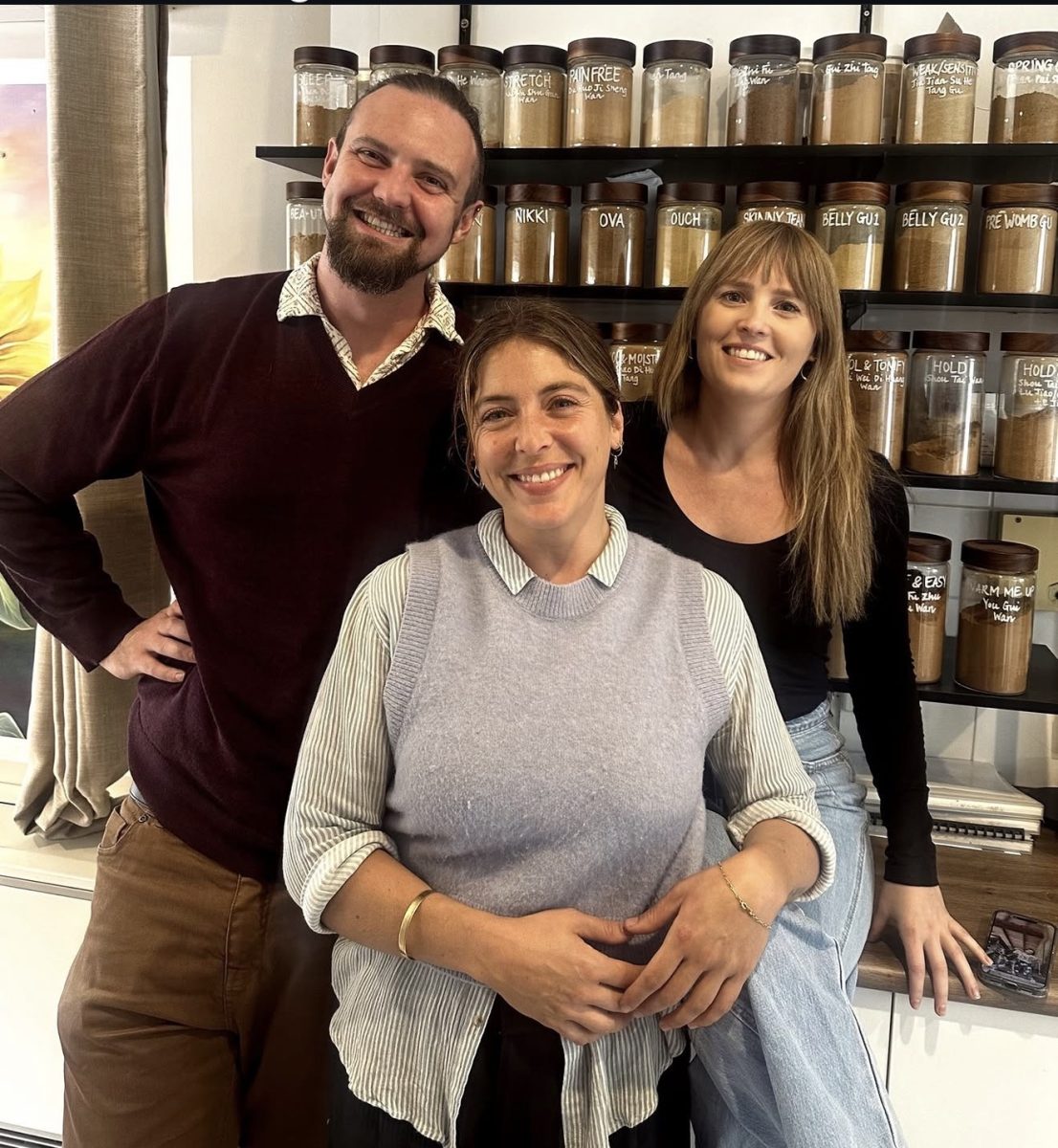 Three people smiling and posing before shelves lined with labelled jars filled with spices or ingredients. Mornington Chinese Medicine